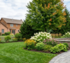 A beautifully landscaped suburban backyard in Amherst, NY, featuring a gray stone paver walkway, white hydrangeas, purple coneflowers, and a wooden pergola over a custom patio.
