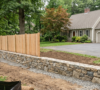 A professionally constructed New England stone retaining wall and wooden privacy fence built on a sloped residential property in Amherst, demonstrating compliance with local town landscape construction codes and setback rules.